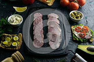 Grey cutting board with salami and ham. Around tomato, souce, olives, lemon, olive oil with spice and sandwiches