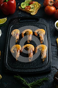 Grey cutting board with shrimp. Around tomato, souce, olives, lemon, olive oil with spice and sandwiches