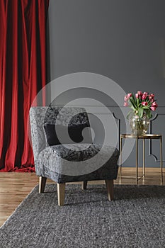 Grey floral chair with black cushion standing on dark carpet in elegant room interior with fresh tulips on gold metal table and re