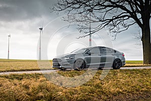 grey Ford Mondeo on a narrow road in a field with wind turbines