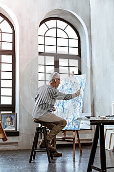 Grey-haired artist painting on canvas in spacious workroom