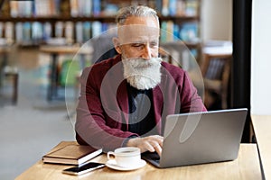 Grey-haired businessman using laptop at cafe, drinking coffee
