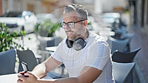 Grey-haired man using smartphone wearing headphones at coffee shop terrace