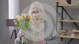 Grey-haired old woman smelling bouquet of flowers looking at camera smiling. Medium shot portrait of happy Caucasian