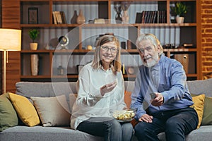 Grey-haired older couple at home choose a television show to watch and switch channels with a TV remote control. elderly mature