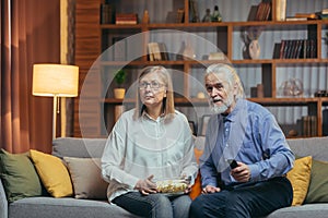 Grey-haired older couple at home choose a television show to watch and switch channels with a TV remote control. elderly mature