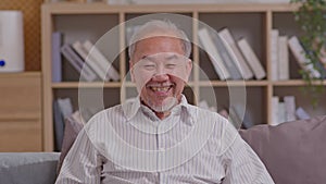 Grey haired senior asian man wearing casual white shirt sitting on couch waving saying hello happy and smiling in video conference