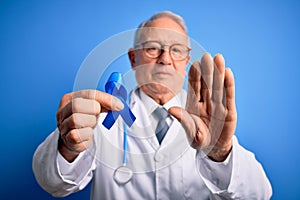 Grey haired senior doctor man holding colon cancer awareness blue ribbon over blue background with open hand doing stop sign with