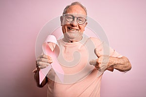 Grey haired senior man holding breast cancer awareness pink ribbon over pink background happy with big smile doing ok sign, thumb