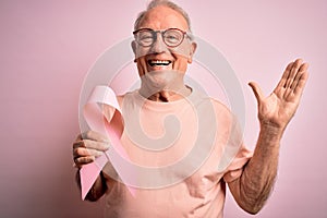 Grey haired senior man holding breast cancer awareness pink ribbon over pink background very happy and excited, winner expression