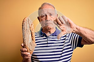 Grey haired senior man holding healthy wholemeal bread over yellow isolated background with angry face, negative sign showing