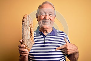 Grey haired senior man holding healthy wholemeal bread over yellow isolated background with surprise face pointing finger to