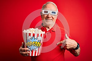 Grey haired senior man wearing 3d movie glasses and eating popcorn over red background with surprise face pointing finger to