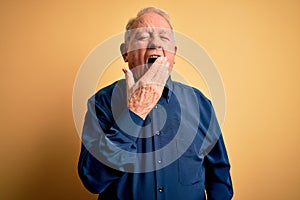 Grey haired senior man wearing casual blue shirt standing over yellow background bored yawning tired covering mouth with hand