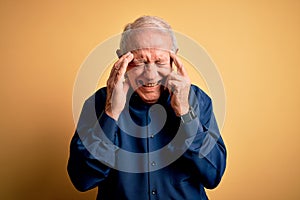 Grey haired senior man wearing casual blue shirt standing over yellow background with hand on headache because stress