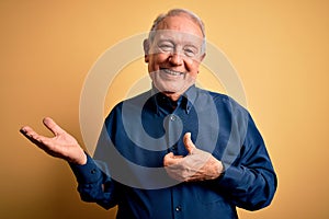 Grey haired senior man wearing casual blue shirt standing over yellow background Showing palm hand and doing ok gesture with