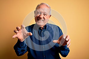 Grey haired senior man wearing casual blue shirt standing over yellow background smiling funny doing claw gesture as cat,