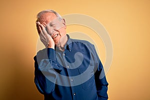 Grey haired senior man wearing casual blue shirt standing over yellow background Yawning tired covering half face, eye and mouth