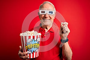 Grey haired senior man wearing 3d movie glasses and eating popcorn over red background with a happy face standing and smiling with