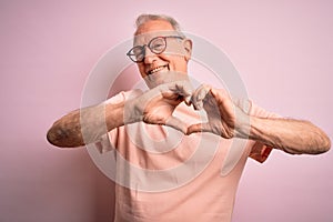 Grey haired senior man wearing glasses standing over pink isolated background smiling in love showing heart symbol and shape with