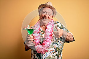 Grey haired senior man wearing summer hat and hawaiian lei drinking a cocktail happy with big smile doing ok sign, thumb up with