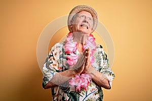 Grey haired senior man wearing summer hat and hawaiian lei over yellow background begging and praying with hands together with