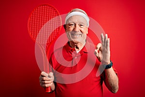 Grey haired senior tennis player man holding racket over red isolated background doing ok sign with fingers, excellent symbol