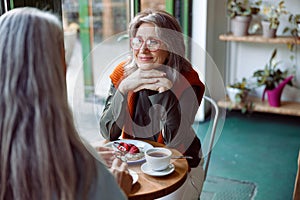 Grey haired woman with glasses looks at friend sitting at small table in cozy cafe