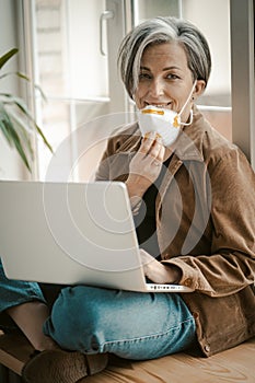 Grey haired woman removes mask affably looking at camera. Caucasian lady uses computer sitting tailor-fashion with