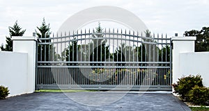 Grey metal wrought iron driveway property entrance gates set in white concrete brick fence, garden trees in background