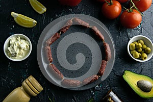 Grey plate with clear space on a black background, around the sausage tomato, souce, olives, lemon, olive oil with spice