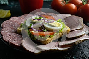 Grey plate with sandwich on a black background, around the salami and ham, tomato, souce, olives, lemon, olive oil with spice