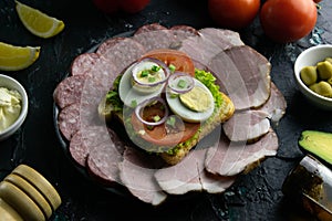 Grey plate with sandwich on a black background, around the salami and ham, tomato, souce, olives, lemon, olive oil with spice