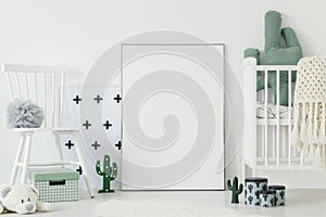 Grey pompom placed on white chair standing in bright baby bedroom interior with cactus shaped decor and empty poster on the floor