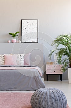 Grey pouf next to bed with cushions in modern bedroom interior with poster and plants. Real photo photo