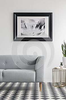 Grey settee on checkered floor in white living room interior with gold table and poster. Real photo