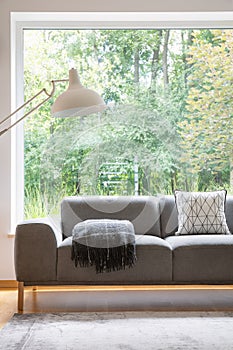 Grey settee with patterned cushion and blanket standing in real photo of sitting room interior with carpet, oversize lamp and big