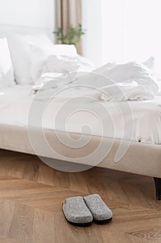 Grey slippers standing near bed with white linens