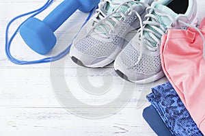 Grey sneakers,  blue dumbbell, jump rope, pink top, leggings on a white wooden background top view