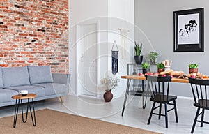 Grey sofa against red brick wall in flat interior with poster and black chairs at dining table. Real photo