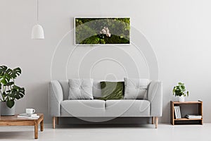 Grey sofa with pillow in real photo of bright sitting room interior with books on wooden shelf, coffee cup on table and garden in