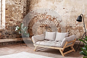 Grey sofa between plant and black lamp in wabi sabi loft interior with red brick wall