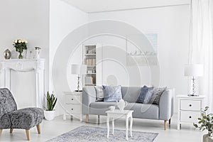 Grey sofa between white cabinets with lamps in flat interior with armchairs and table on carpet. Real photo