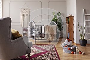 Grey and stylish baby room interior with wooden crib and handmade macrame on the empty wall