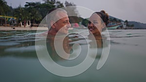 Greyhaired man and asian girlfriend look at each other in the water