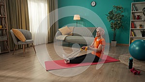 Greyhaired woman aged exercising with an elastic sports band. An elderly woman is sitting on the floor, the expander