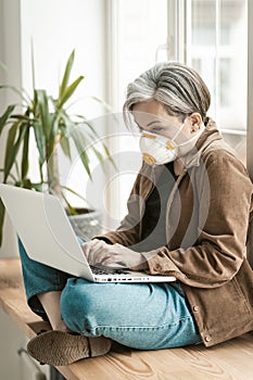 Greying businesswoman works laptop from home. Aged woman wearing protective mask uses mobile internet on computer