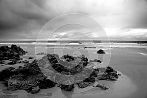 Greyscale of the beach surrounded by rocks and a wavy sea under a cloudy sky at daytime