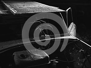 Greyscale closeup of an old car with dismantled parts in an old creepy room