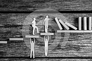 Greyscale image of small paper people holding up falling dominos while standing on wooden steps